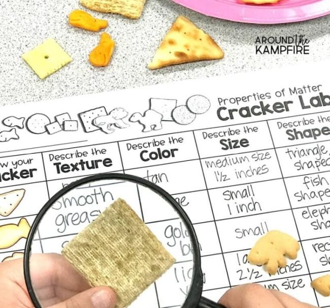 Child's hand holding magnifying glass over a cracker, with worksheet labeled Cracker Lab in the background-science projects for 2nd graders