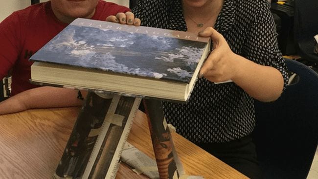 Students balancing a textbook on top of a pyramid of rolled up newspaper