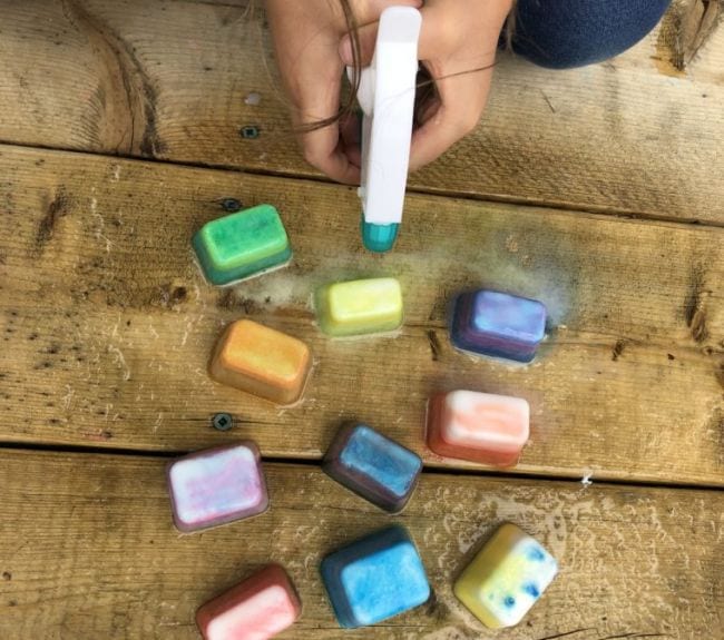 Child's hands holding a spray bottle over colorful ice cubes on a wood surface as an example of kindergarten science experiments