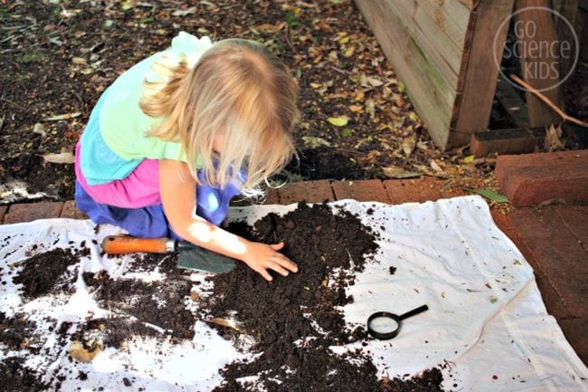 Child examining dirt on a white tarp with a magnifying glass as an example of kindergarten science experiments