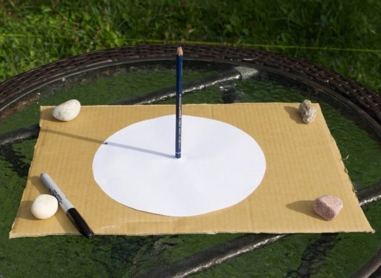 A large piece of cardboard has a white circle in the center with a pencil standing upright in the middle of the circle. Rocks are on all four corners holding it down. (easy science experiments)