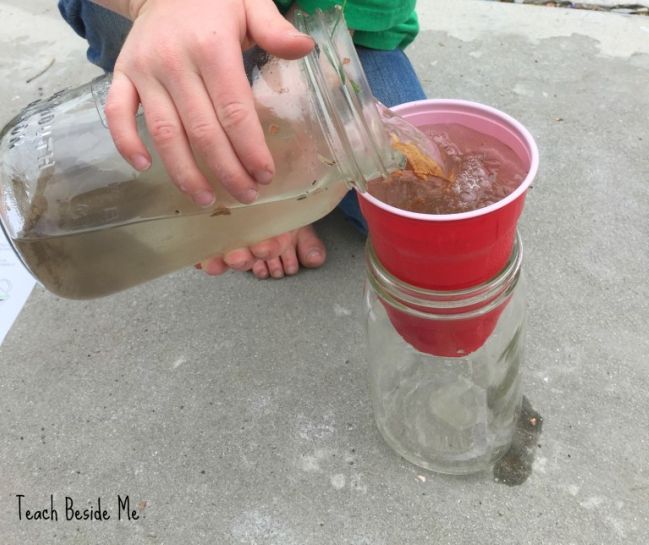 Child pouring dirty water into a cup sitting on top of a large mason jar in this example of 5th grade science