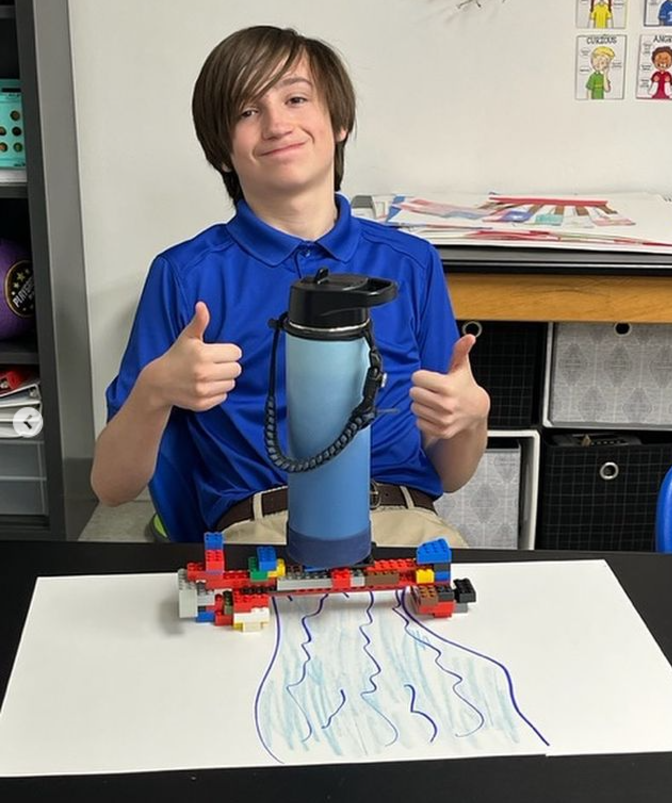 A middle school boy is seen in front of a LEGO bridge he built with a water bottle sitting on top of it in this example of 6th grade science fair projects.