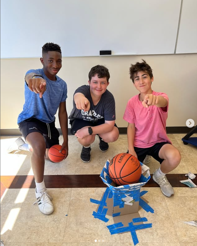 Three middle school aged boys kneel in front of a structure made of cardboard and tape that is holding a basketball in this example of 6th grade science fair projects.