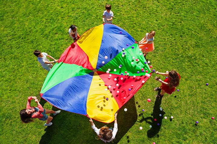 Students stand around a large parachute with small balls bouncing on the top of it as an example of elementary PE games
