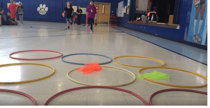 Students stand in the background. In the foreground are several hula hoops laid out on the floor as an example of elementary PE games