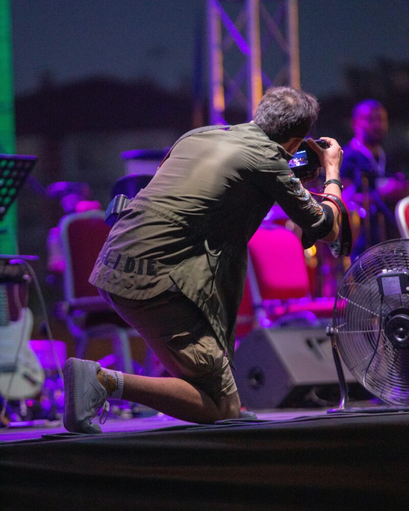 A man is seen taking a photograph from behind of a stage with musicians on it in this example of music careers.