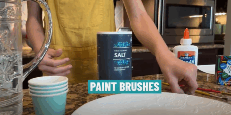 SaltPainting_Ingredients A child is seen from the neck down standing in front of a table that has cups, a plate, a pitcher, a container of salt, Elmer's glue, or food coloring.