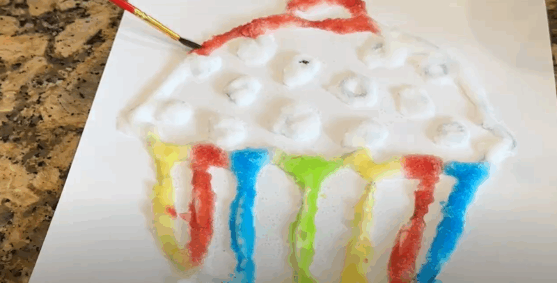 SaltPainting_Step5 A cupcake coloring page has glue and salt spread on it. A paintbrush is shown spreading color onto it.