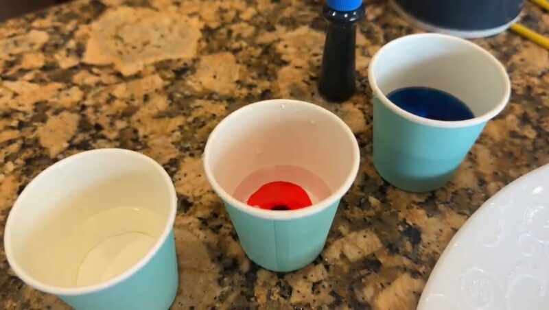 SaltPainting_foodcoloring Several small paper cups are shown on a counter top filled with water. A drop of food coloring has been added into the cups in this second step of salt painting.