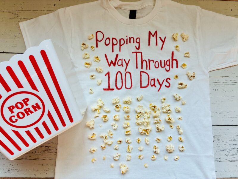 a white shirt with the words popping my way through 100 days and 100 pieces of popcorn and a red and white popcorn container